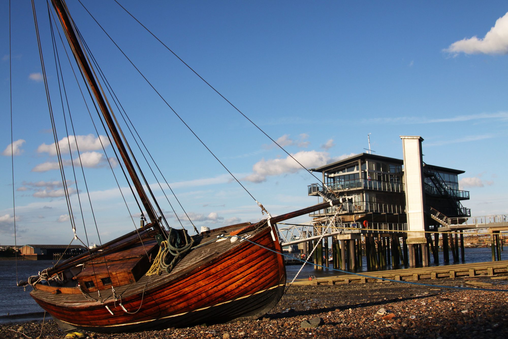A wooden sailing barge on the foreshore at low tide with the clubhouse on its piles behind under a blue sky