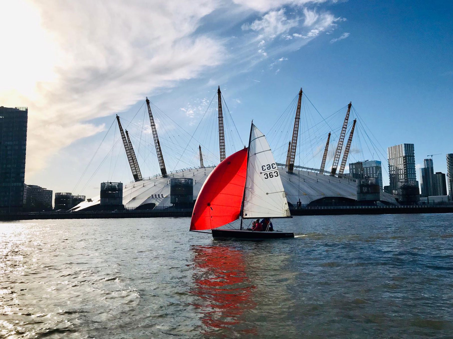 A dinghy with a red spinnaker racing on the Thames in front of the O2