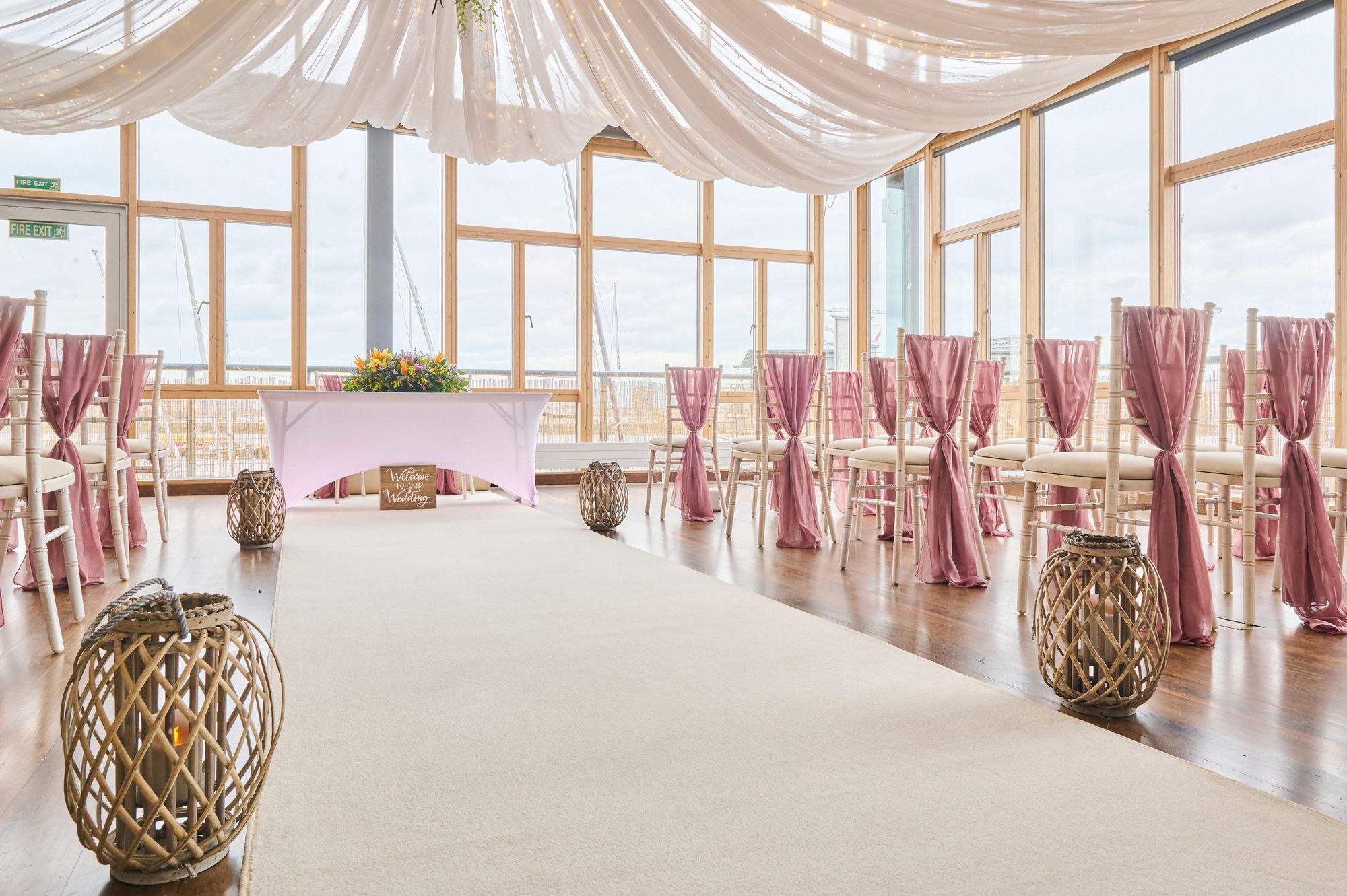 The River Rooms set for a wedding ceremony: white aisle carpet, limewash chairs with dusky pink sashes, wicker lanterns and a draped ceiling
