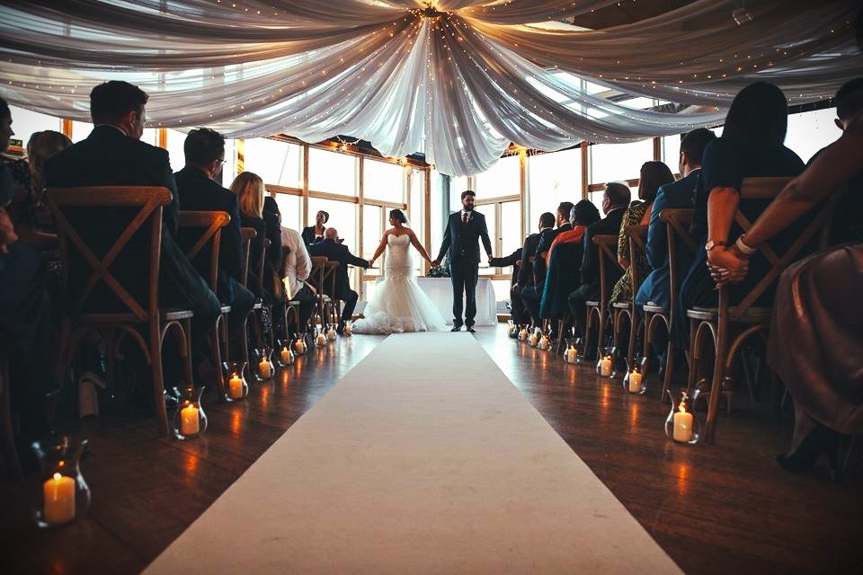 A newly married couple walk hand in hand back down the aisle between candle lanterns and their guests in the River Rooms
