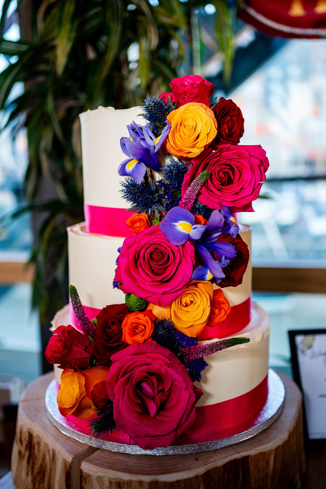 A three tier white wedding cake with a red ribbon and cascading red, orange and purple flowers