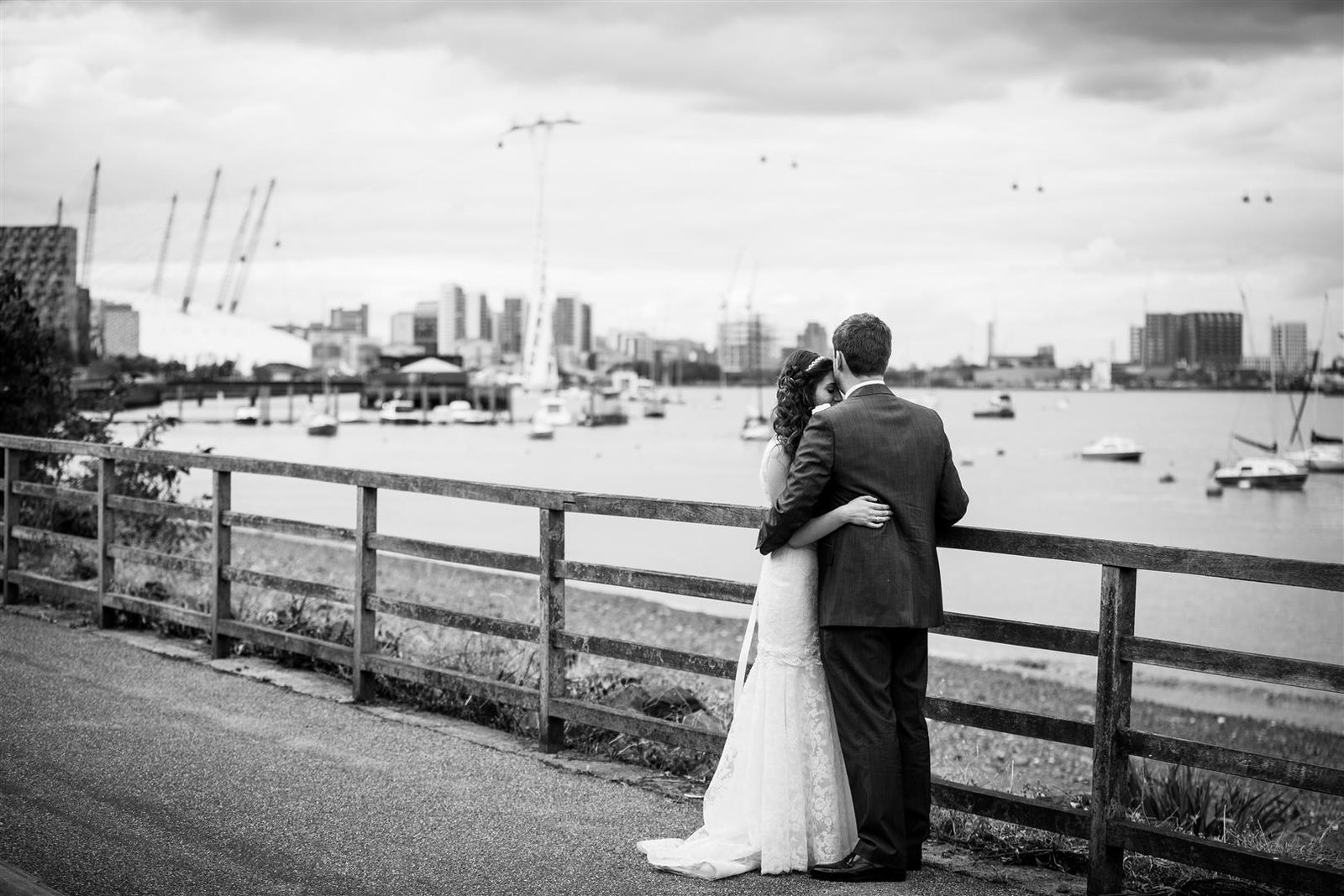 Black and white photograph of a bride and groom embracing at the railing on the riverside path with the Thames and Canary Wharf behind