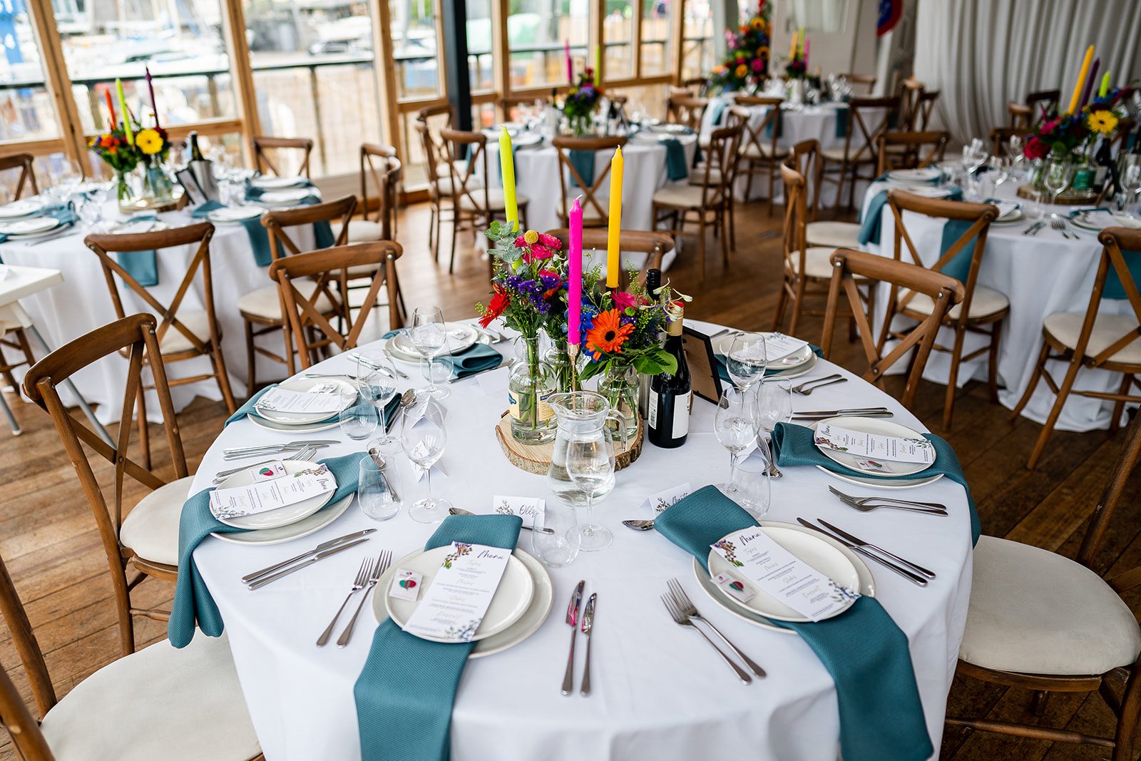 A round table laid with teal napkins, coloured candles and bright flowers, crossback chairs and the river beyond