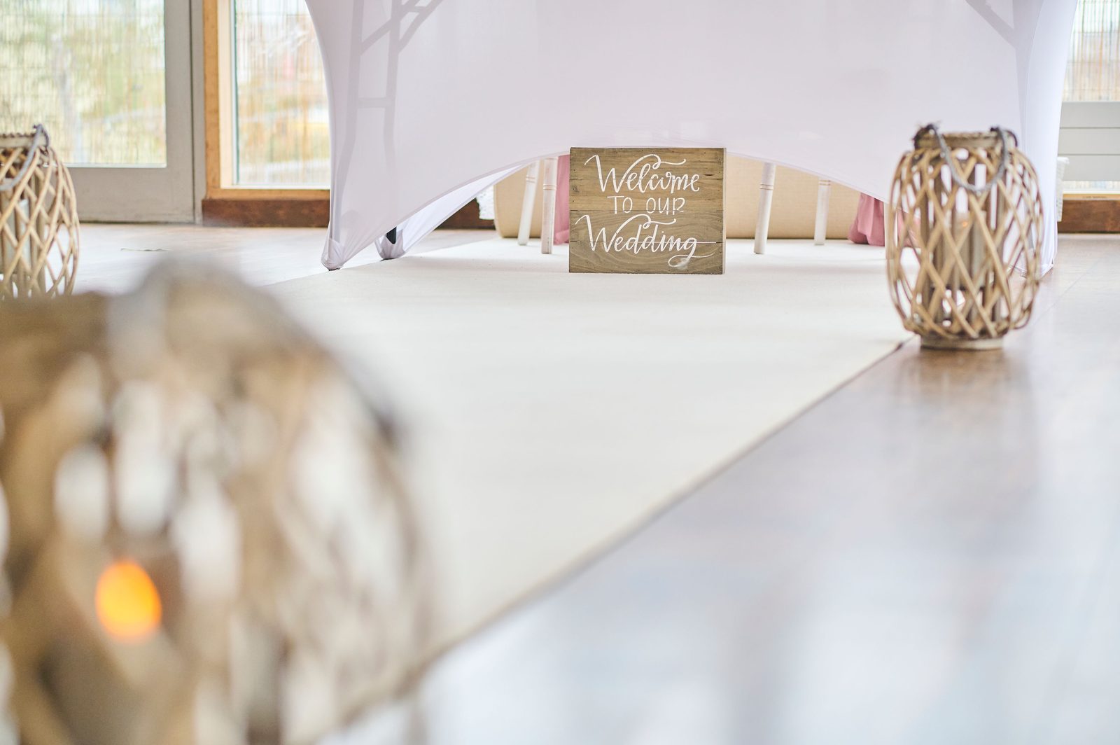 A Welcome to our Wedding sign on the signing table at the end of the white aisle, wicker lanterns either side